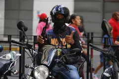 Um homem em uma moto armado e com o rosto coberto por uma balaclava, dando um joinha para a câmera