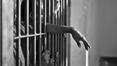 ከእስር ቤት በር የብረት ፍርግርግ ጀርባ እጁ ወጥቶ የሚታይ ሰው ጥቀር እና ነጭ ፎቶ