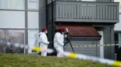 دو تن از افراد فنی طب عدلی که لباس‌های سفید و کلاه سرخ و ماسک آبی دارند، در حال تحقیق در مقابل منزل هستند.