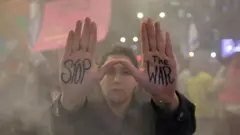 Une femme manifestant contre le gouvernement et soutenant les otages du 7 octobre lève les deux mains, avec les mots « Stop the War » écrits sur ses paumes, tandis que des manifestants avec des banderoles se tiennent à l'arrière-plan