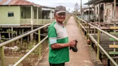 Homem usando um boné e uma camisa verde olha para a câmera. Ele segura uma sacola preta nas mãos Atrás dele há casas sob o rio