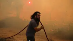 Человек тащит пожарный шланг на фоне оранжевого от дыма пейзажа