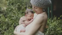Жінка в сірій вʼязаній шапці тримає немовля на руках