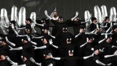 Alok se apresenta ao fundo do palco, cercado por dezenas de performers vestidos de preto, com braços prateados e máscaras espelhadas que ocultam seus rostos. Os dançarinos executam uma coreografia sincronizada com os braços erguidos em diferentes direções.