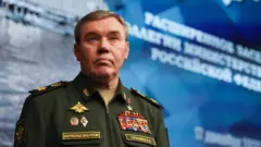 Чоловік у формі російського генерала, з оголеною головою, стоїть на блакитному тлі з білими літерами