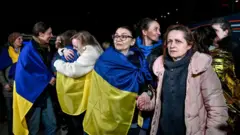 Varias mujeres envueltas en banderas ucranianas azules y amarillas. Dos mujeres se abrazan al fondo y otras sonríen. Liudmyla mira hacia su derecha mientras sostiene la mano de una mujer con abrigo rosa situada a su izquierda.