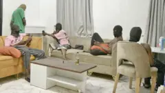 Foto do interior do Hustle Kingdom. Quatro homens sentados em sofás segurando celulares. Todos usam balaclavas escondendo o rosto – dois usam camisetas rosas, outro usa um colete branco e outro está sem camisa.