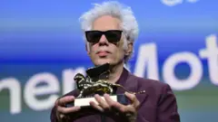 Седой Джим Джармуш с темных очках и пурпурном пиджаке держит перед собой в руках статуэтку в виде золотого льва