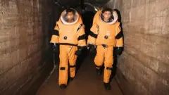 Duas pessoas em trajes espaciais laranja atravessando um túnel de concreto