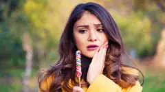 una mujer sostiene un dulce mientras lleva su mano izquierda a la mejilla con rostro de preocupación. Tiene pelo largo marrón y lleva un abrigo amarillo. 