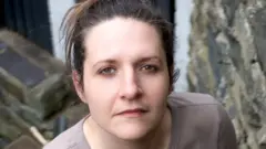 Ruth O'Grady olhando para a câmera, ao lado de uma parede de pedras, vestindo uma blusa de mangas longas