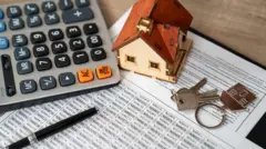 Foto de uma calculadora sobre uma planilha com um molho de chaves, uma caneta e uma miniatura de casa.
