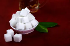 مكعبات سكر لونها أبيض داخل صحن صغير بتدرجات الألوان الزهري والأبيض وبجانبه ورقتا شجر لونهما أخضر وثلاث مكعبات سكر بيضاء من الجانب الآخر وقرب الصحن كأس زجاجي يبدو أنه يحتوي على الشاي. الأرضية يميل لونها إلى الخمري. 
