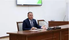 Собиқ Қумқўрғон ҳокими Анвар Норамамтов мажлисда стол қаршисида ўтириб, микрафонга гапираяпти. У расмда тўқ кўк ранг костюм шимда, оқ куйлак, кўк гакстук таққан.