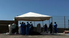 Personal médico alrededor de cadáveres sobre el suelo cubiertos con bolsas de plástico. Los trabajadores llevan batas azules o blancas.