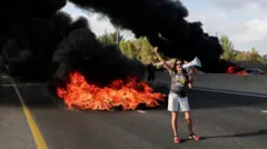 Женщина с мегафоном на шоссе, позади нее горящий мусор и клубы черного дыма