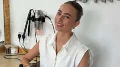 Mulher de camiseta branca com cabelos presos, sorrindo para a câmera, com um secador de cabelos ao fundo