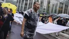 黒い衣服とビニール製の上着を着た男性が、声を張り上げながら、大きな横断幕の端を持ち、行進している。周囲には傘を差しながら同じように横断幕を持って歩いている人々がいる