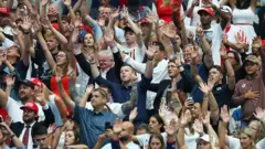 Um grupo de pessoas de diferentes idades e raças levanta os braços em louvor durante o evento em memória de Charlie Kirk.