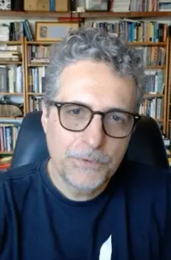 Kleber Mendonça está sentado em uma cadeira. Atrás dele há uma estante com vários livros