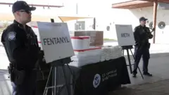 Mesa repleta de pacotes de pó branco e dois cartazes dizendo 'fentanil' e 'meta', vigiada por dois homens de uniforme e boné