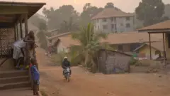 A motorcycle drives down a dirt road in Sierra Leone as children watch from the balcony.