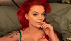 Uma mulher ruiva com o dedo na boca. Ela está deitada em uma cama fazendo uma selfie.