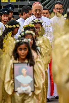 දැරියන් පිරිසක් පාස්කු ප්‍රහාරයෙන් මිය ගිය අයගේ ෆොටෝ රැගෙන පෙළට ගමන් කරන අතර කාදිනල් හිමියන් ඇතුළු පිරිසක් පසුපසින් සිටී.