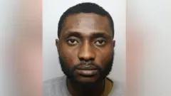 Mugshot of Chiemka Okoronta. Im wear grey sweatshirt and get short black hair and beard. 