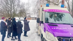 Cinco mulheres usando roupas de inverno estão em frente a uma ambulância.