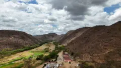 Povoado localizado em vale com margens esverdeadas; em volta, há morros com paisagem semiárida