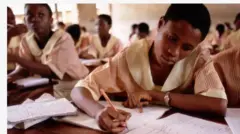 Aworan awọn akẹkọọ to n kọwe ni kilaasi. Obinrin ni awọn akẹkọọ naa, wọn wọ aṣọ ileewe alawọ erupẹ, akẹkọọ to hanju mu gègé lọwọ, o n kọwe lọwọ, iwe meji wa niwaju rẹ, awọn iwe kan si tun wa lorii tabili naa pẹlu