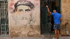 مردی از پشت سر جلوی در خانه‌ای ایستاده، دیوارنگار کنار در تصویر محو چه‌گه‌وارا است