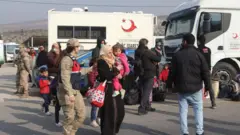 Göç İdaresi'ne ait araçların önünde çocuğunu taşıyan bir kadın, yanlarında görevliler var 