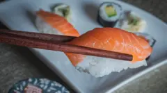 A salmon nigiri held with chopsticks, with a plate of a selection of sushi in the background.