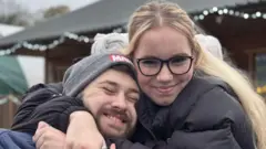Megan Dixon abraçando o namorado. Megan é loira e está usando óculos com armação preta e um casaco preto. Oliver usa uma touca cinza e barba e está de olhos fechados sorrindo.