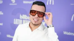 Yeison Jiménez posa ante la cámara llevándose la mano izquierda a las gafas delante de un fondo morado.