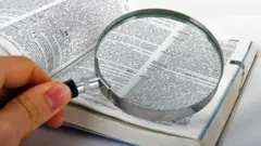 A hand holds a magnifying glass over an open page of a dictionary.
