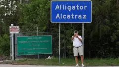 Homem de camisa branca leva as mãos à cabeça junto a uma placa indicativa do 'Alcatraz dos jacarés', na Flórida (EUA)