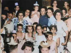 Crianças e adultos posando para foto. Algumas crianças usam chapéus festivos e todas estão vestidas com roupas de festa. Alina Fernández está no alto da foto com um chapéu cônico de festa e um vestido rosa-claro.