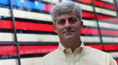 Um homem branco de cabelos grisalhos em frente a uma bandeira dos EUA