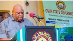 Gomina ipinle Osun, Ọmọọba Ademola Adeleke n sọrọ lọwọ ninu aworan yii. Aṣọ alawọ eeru lo wọ, ẹrọ gbohungbohun si wa niwaju rẹ.