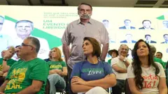 Michelle Bolsonaro em frente a uma imagem de Bolsonaro com a boca tampada com a mensagem "falem por mim"