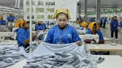 A women wearing a dark blue shirt and a yellow headcloth in a garment factory in Cambodia. She is tending to fabric on a table. Behind her are rows of tables with other similarly dressed women.