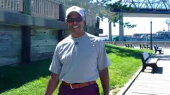Tom Teixeira, con una camiseta gris y pantalones cortos, camina junto al río en Fall River, con el puente Braga de fondo.