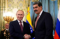 Vladimir Putin cumprimenta Nicolás Maduro. Ambos usam terno escuro e gravata, com bandeiras da Rússia e da Venezuela ao fundo.