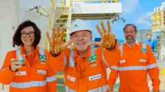 Lula e outros dois representantes da Petrobras, todos vestindo uniformes laranjas, posam sorrindo em uma plataforma offshore; Lula aparece ao centro, com as mãos erguidas, acompanhado de Magda Chambriard à esquerda e outro executivo à direita.