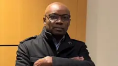 Charles Onana ahagaze imbere y'umuryango w'urukiko i Paris mu Bufaransa, asobekeranyije amaboko, yambaye ikoti ry'umukara n'indorerwamo. Ifoto yo ku itariki ya 9/12/2024