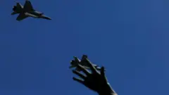 На тлі чистого синього неба летять два військові літаки. У нижній частині кадру видно підняту догори розкриту руку, силует якої частково перекриває один із літаків.