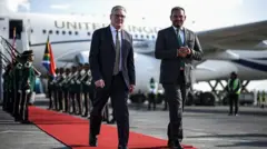 Стармер идет по красной дорожке от самолета в сопровождении представителя ЮАР